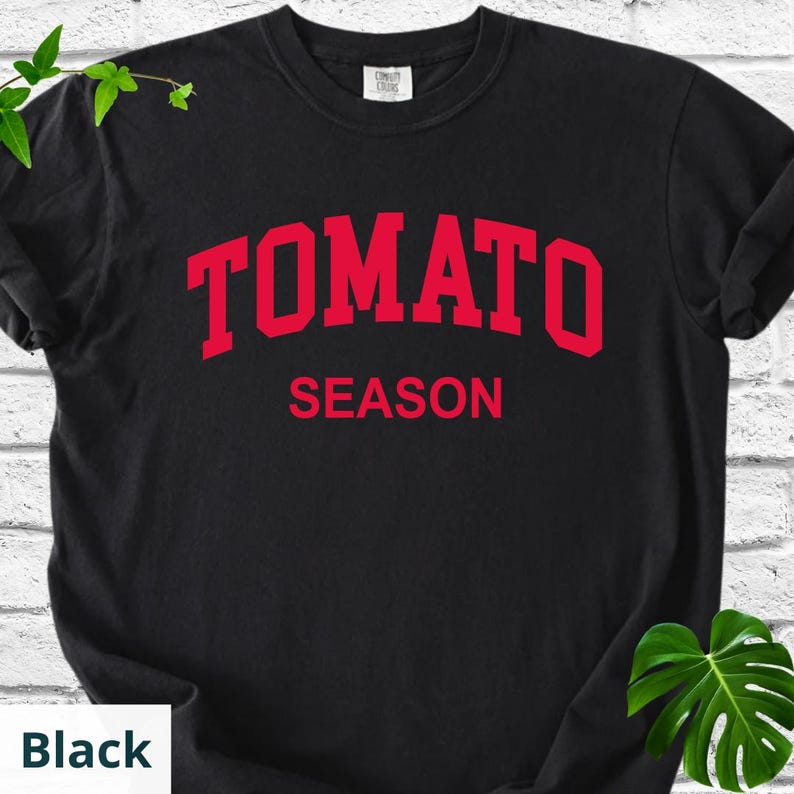 K&ouml;nnte beinhalten: Schwarzes T-Shirt mit dem roten Schriftzug "TOMATO SEASON" in bogenf&ouml;rmigen Buchstaben. Das T-Shirt hat einen Rundhalsausschnitt und scheint aus einem weichen Material zu sein. Der Text ist mittig auf der Vorderseite des Shirts platziert.