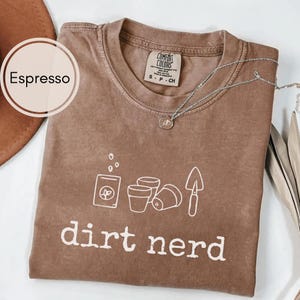 May include: Espresso-coloured t-shirt with a white graphic of gardening tools and the words "dirt nerd." The shirt has a crew neck and short sleeves. A silver necklace with a small pendant is draped across the shirt.