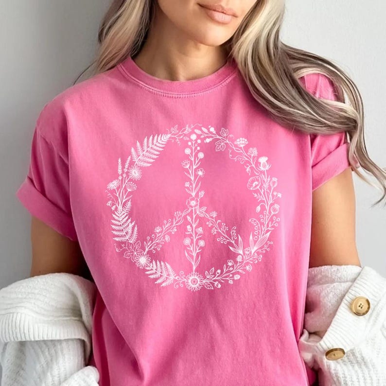May include: A bright pink short-sleeve t-shirt featuring a large, white, circular graphic print on the chest. The graphic is composed of various delicate botanical illustrations, including ferns and wildflowers, arranged to form a peace sign symbol.