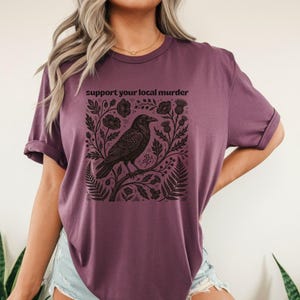 May include: A plum-colored short-sleeve t-shirt featuring a black graphic print of a crow perched on a branch surrounded by botanical elements like ferns and thistles. The text "support your local murder" is printed in black above the illustration.