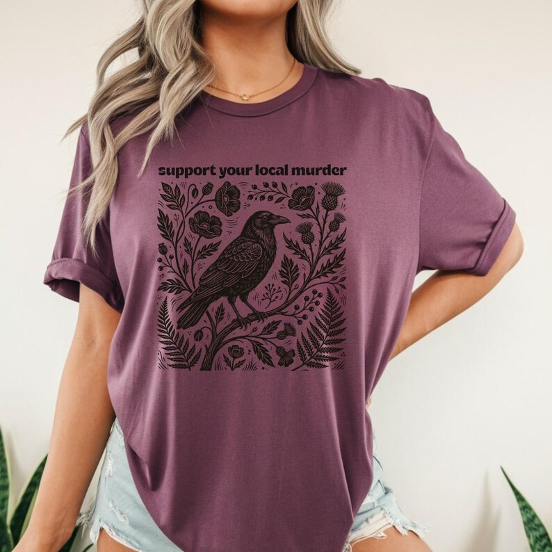 May include: A plum-colored short-sleeve t-shirt featuring a black graphic print of a crow perched on a branch surrounded by botanical elements like ferns and thistles. The text "support your local murder" is printed in black above the illustration.