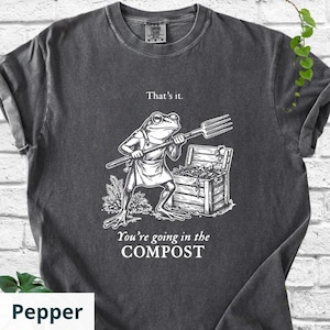 May include: Pepper-colored t-shirt with a humorous graphic. The design features a frog wearing an apron and holding a pitchfork, standing in front of a wooden compost bin. Text above reads "That's it." and below, "You're going in the COMPOST."