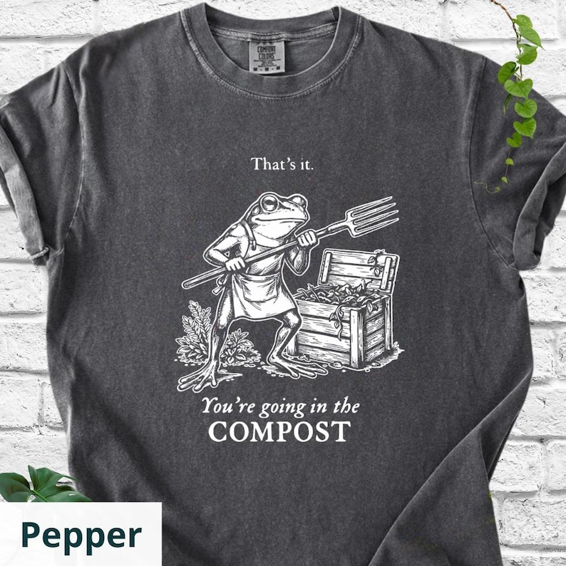 May include: Pepper-colored t-shirt with a humorous graphic. The design features a frog wearing an apron and holding a pitchfork, standing in front of a wooden compost bin. Text above reads "That's it." and below, "You're going in the COMPOST."