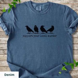 May include: A blue short-sleeved t-shirt featuring a black graphic of four crows perched in a row. Below the birds, the text reads "support your local murder" in a typewriter-style font. The shirt has a crew neckline and a washed-out appearance.