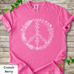 May include: A bright pink short-sleeve t-shirt featuring a white graphic of a peace sign composed of various botanical elements, including ferns, wildflowers, and leaves. The shirt has a crew neckline and a Comfort Colors brand label at the neck.