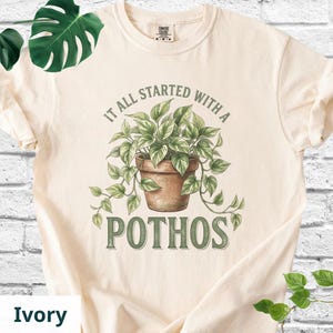 May include: An ivory short-sleeve t-shirt featuring a graphic of a green pothos plant in a brown terracotta pot. The text above the plant reads IT ALL STARTED WITH A and the text below the plant reads POTHOS in a dark green serif font.