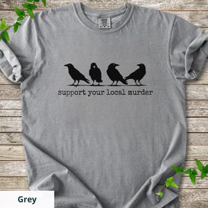 May include: A grey short-sleeved t-shirt featuring a black graphic of four crows standing in a row. Below the birds, the text reads "support your local murder" in a typewriter-style font. The shirt has a crew neckline and a standard fit.