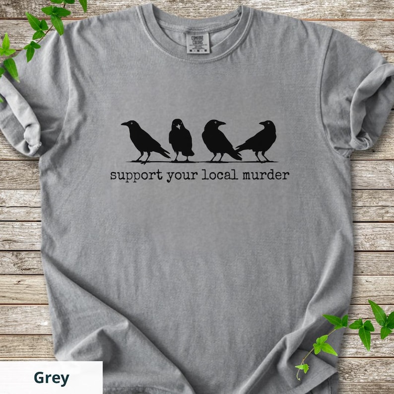 May include: A grey short-sleeved t-shirt featuring a black graphic of four crows standing in a row. Below the birds, the text reads "support your local murder" in a typewriter-style font. The shirt has a crew neckline and a standard fit.