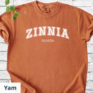 May include: A rust-coloured t-shirt featuring the word "ZINNIA" in white, curved lettering above the word "SEASON". The shirt has a crew neck and short sleeves.
