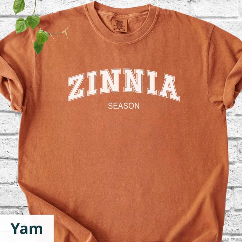 May include: A rust-coloured t-shirt featuring the word "ZINNIA" in white, curved lettering above the word "SEASON". The shirt has a crew neck and short sleeves.