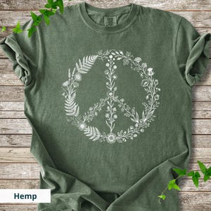 May include: A forest green short-sleeve t-shirt featuring a white graphic print of a peace sign composed of various botanical elements, including ferns, wildflowers, and leafy stems arranged in a circular pattern on the front of the garment.