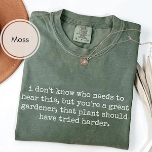 May include: A moss green t-shirt with the text "i don't know who needs to hear this, but you're a great gardener, that plant should have tried harder." A silver necklace with a pendant is on the shirt.
