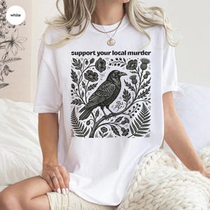 May include: A white short-sleeve t-shirt featuring a black graphic print of a crow perched on a branch surrounded by botanical elements like flowers and ferns. The text "support your local murder" is printed in black above the crow illustration.