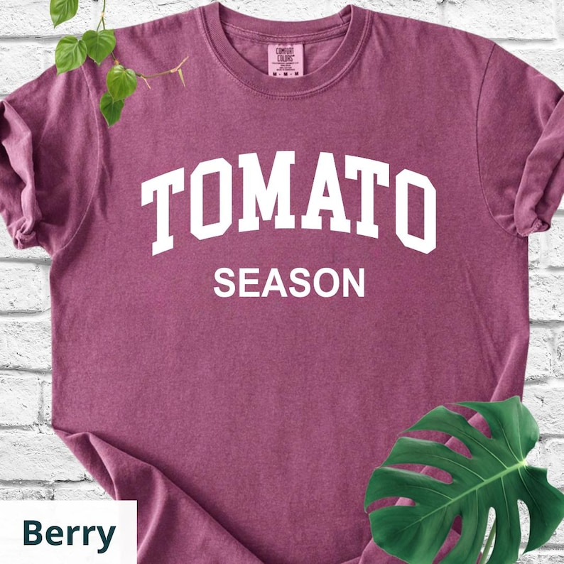 K&ouml;nnte beinhalten: Beerenfarbenes T-Shirt mit den Worten "TOMATO SEASON" in wei&szlig;en Blockbuchstaben. Das Shirt hat einen Rundhalsausschnitt und kurze &Auml;rmel. Das Shirt ist aus einem weichen Material. Das Wort "Berry" steht in einem wei&szlig;en Feld unten links.