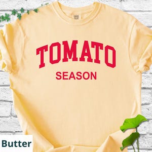 K&ouml;nnte beinhalten: Hellgelbes T-Shirt mit dem roten Schriftzug "TOMATO SEASON". Das T-Shirt hat kurze &Auml;rmel und einen Rundhalsausschnitt. Der Text ist mittig auf der Vorderseite des Shirts platziert.
