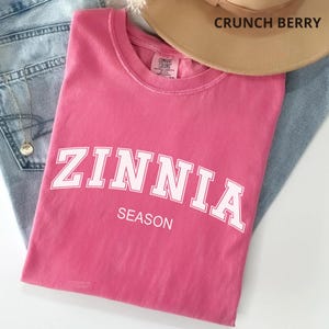 May include: A pink short-sleeved t-shirt featuring the white text ZINNIA SEASON in a collegiate font across the chest. The shirt has a crew neckline and a Comfort Colors brand label on the inside collar. The fabric has a soft, washed appearance.