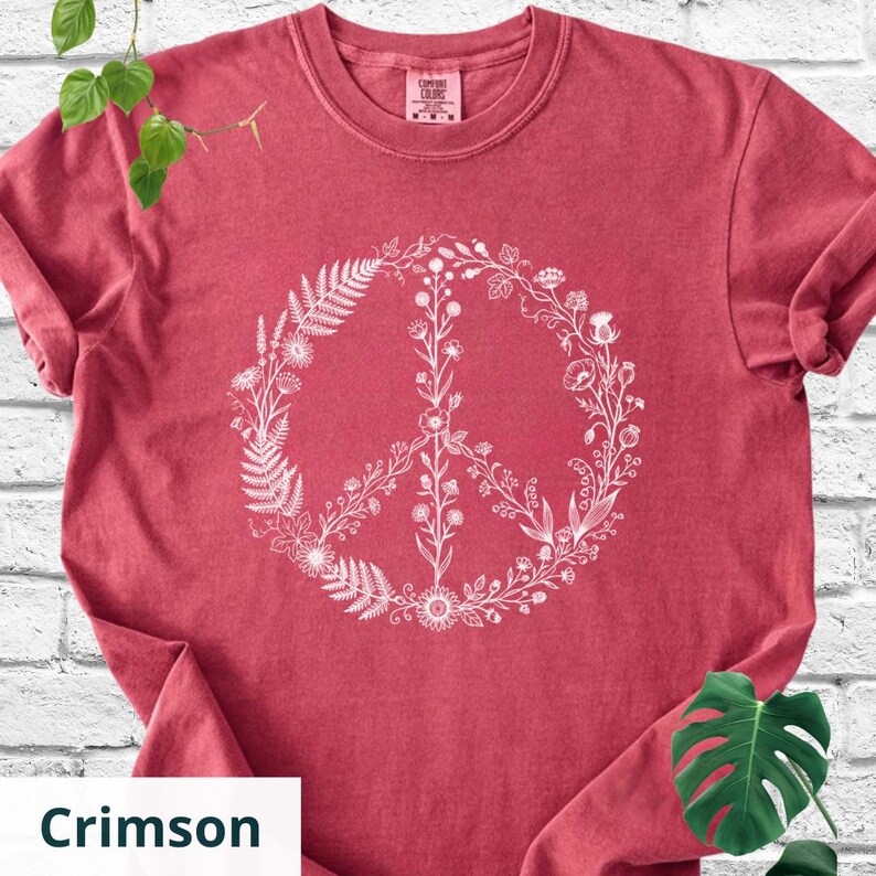 May include: A crimson short-sleeve t-shirt featuring a white graphic print of a peace sign composed of various botanical elements, including ferns, wildflowers, and leafy stems. The shirt has a crew neckline and a Comfort Colors brand label.
