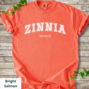 May include: A bright salmon-coloured short-sleeve t-shirt featuring the white text ZINNIA arched across the chest with the word SEASON printed in a smaller white font directly below. The shirt has a crew neckline and a visible brand tag.