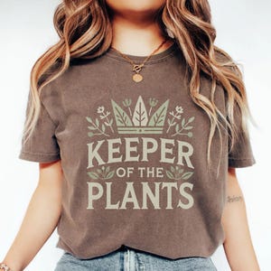 May include: A brown t-shirt with the text "KEEPER OF THE PLANTS" in a light green font. The text is surrounded by a crown and floral design. The shirt has short sleeves and a crew neck.