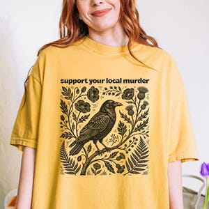 May include: A mustard yellow short-sleeve t-shirt featuring a black graphic print of a crow perched on a branch surrounded by flowers and foliage. The text "support your local murder" is printed in black above the central crow illustration.