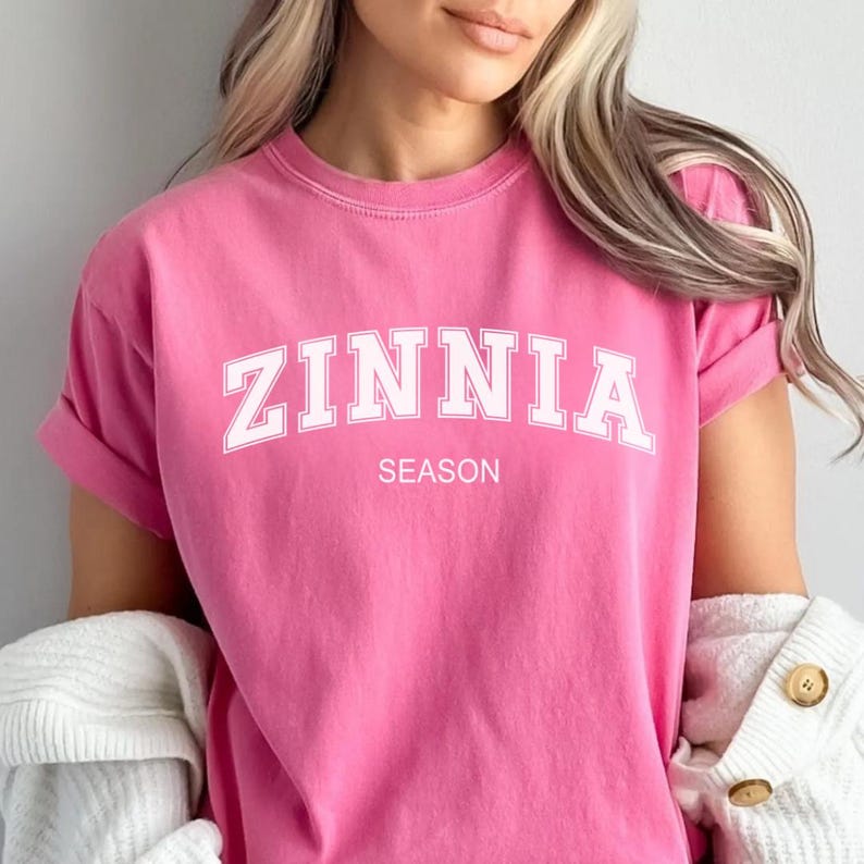 May include: A vibrant pink t-shirt featuring the word "ZINNIA" in white, curved text, and the word "SEASON" underneath. The shirt has rolled sleeves and is made of a soft fabric. A white cardigan is draped over the shoulders.