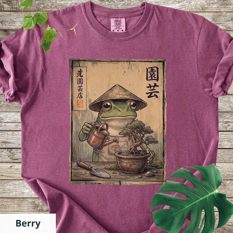 May include: Berry-colored t-shirt featuring a frog wearing a conical hat and watering a bonsai tree. The frog is depicted in a vintage style, with Japanese text and imagery. The shirt has a classic crew neck and short sleeves.
