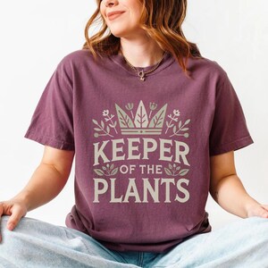 May include: A maroon t-shirt with the text "KEEPER OF THE PLANTS" in a light green font. The text is surrounded by a crown and floral design. The shirt has short sleeves and a relaxed fit.