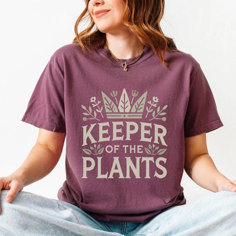 May include: A maroon t-shirt with the text "KEEPER OF THE PLANTS" in a light green font. The text is surrounded by a crown and floral design. The shirt has short sleeves and a relaxed fit.