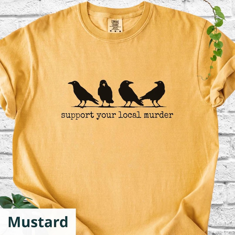 May include: A mustard yellow crew neck t-shirt featuring a black graphic of four crows standing in a row. Below the birds, the text reads "support your local murder" in a typewriter-style font. The shirt has a Comfort Colors brand label.