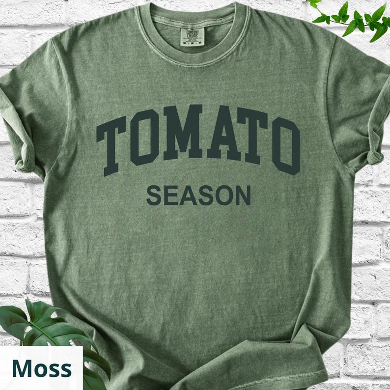 K&ouml;nnte beinhalten: Ein moosgr&uuml;nes T-Shirt mit dem Aufdruck "TOMATO SEASON" in dunkelgr&uuml;nen Blockbuchstaben. Das Shirt hat einen Rundhalsausschnitt und kurze &Auml;rmel. Der Stoff wirkt weich und leicht verblichen.