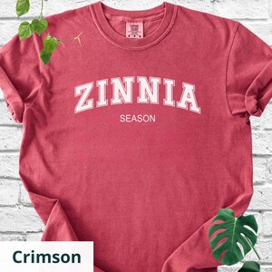 May include: Crimson t-shirt featuring the word "ZINNIA" in white arched lettering above "SEASON". The short-sleeved, crew-neck shirt is made of a soft material. The shirt's colour is described as crimson.