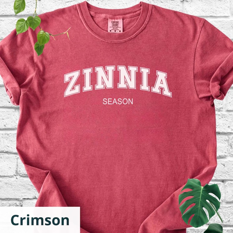 May include: Crimson t-shirt featuring the word "ZINNIA" in white arched lettering above "SEASON". The short-sleeved, crew-neck shirt is made of a soft material. The shirt's colour is described as crimson.