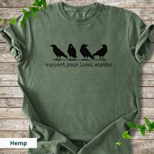 May include: An olive green t-shirt featuring a black graphic of four crows standing in a row. Below the birds, the text reads "support your local murder" in a typewriter-style font. The shirt has a crew neckline and short sleeves.