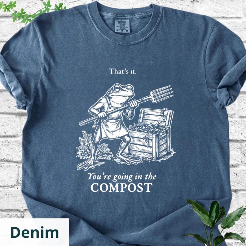 May include: Denim blue t-shirt with a humorous graphic of a frog holding a pitchfork, standing next to a wooden compost bin. The text reads "That's it. You're going in the COMPOST."