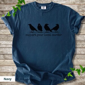 May include: A navy blue short-sleeve t-shirt featuring a black graphic of four crows standing in a row. Below the birds, the text reads "support your local murder" in a simple, dark, typewriter-style font. The shirt has a crew neckline.