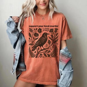 May include: A burnt orange short-sleeve t-shirt featuring a black graphic print of a crow perched on a branch surrounded by flowers and foliage. The text "support your local murder" is printed in black above the crow illustration.