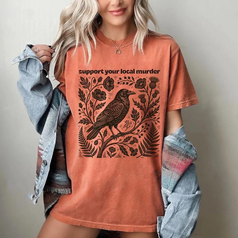 May include: A burnt orange short-sleeve t-shirt featuring a black graphic print of a crow perched on a branch surrounded by flowers and foliage. The text "support your local murder" is printed in black above the crow illustration.