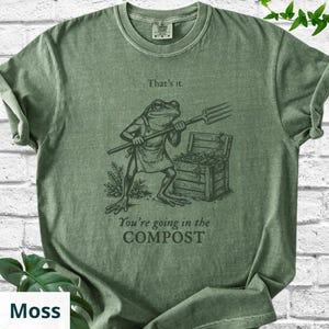 May include: Moss green t-shirt with a graphic of a frog holding a pitchfork, standing near a compost bin. Text on the shirt reads "That's it." and "You're going in the COMPOST."