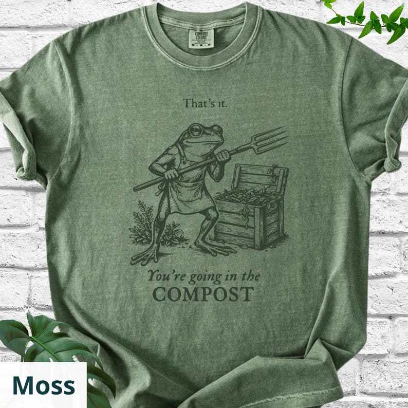May include: Moss green t-shirt with a graphic of a frog holding a pitchfork, standing near a compost bin. Text on the shirt reads "That's it." and "You're going in the COMPOST."