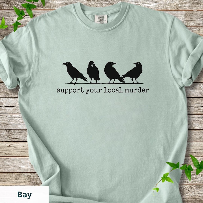 May include: A light sage green short-sleeve t-shirt featuring a black graphic of four crows standing in a row. Below the birds, the text reads "support your local murder" in a typewriter-style font. The shirt has a crew neckline and a standard fit.