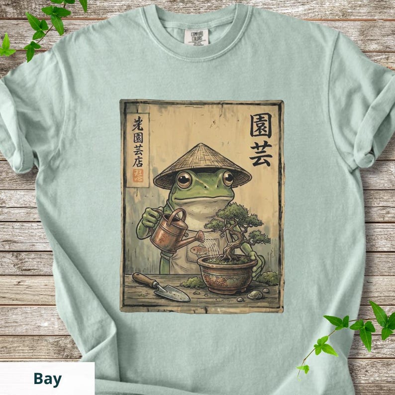 May include: Light blue t-shirt featuring a cartoon frog wearing a conical hat, watering a bonsai tree. The frog is depicted with a watering can and a small shovel. Japanese text is visible on the design.