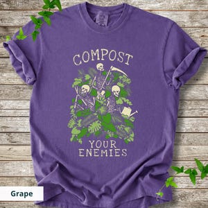 Peut inclure: Un t-shirt violet avec les mots "COMPOST YOUR ENEMIES" dans une police vieillie. Le graphisme repr&eacute;sente des squelettes jardinant parmi un feuillage vert. Le t-shirt est de couleur raisin.
