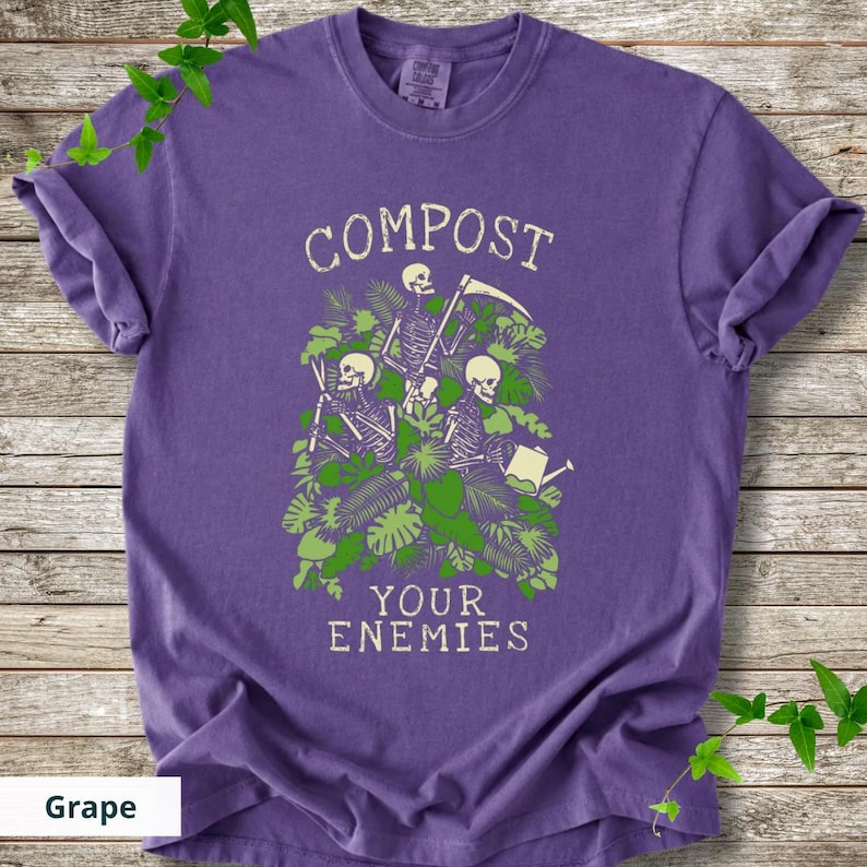 Peut inclure: Un t-shirt violet avec les mots "COMPOST YOUR ENEMIES" dans une police vieillie. Le graphisme repr&eacute;sente des squelettes jardinant parmi un feuillage vert. Le t-shirt est de couleur raisin.