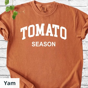 K&ouml;nnte beinhalten: Ein rostfarbenes T-Shirt mit dem Text "TOMATO SEASON" in gro&szlig;en, wei&szlig;en Buchstaben. Das Shirt hat einen Rundhalsausschnitt und kurze &Auml;rmel. Der Stoff wirkt weich und leicht verblichen.