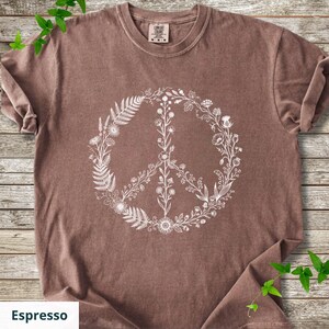 May include: A brown short-sleeved t-shirt featuring a white graphic print of a peace sign composed of various botanical elements, including ferns, wildflowers, and leaves. The shirt has a crew neckline and a Comfort Colors brand label at the neck.