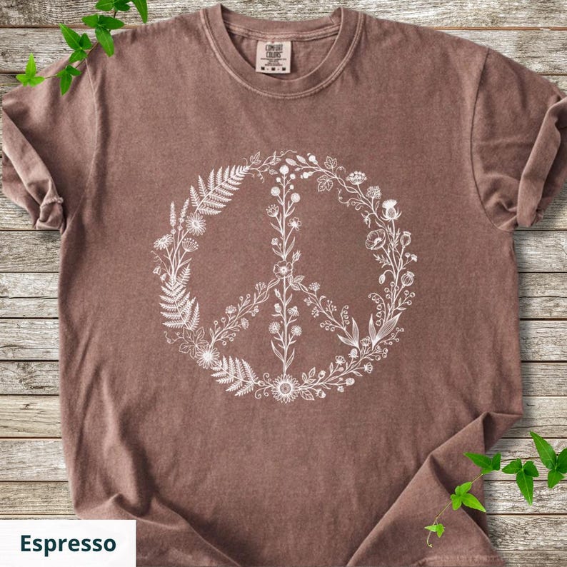 May include: A brown short-sleeved t-shirt featuring a white graphic print of a peace sign composed of various botanical elements, including ferns, wildflowers, and leaves. The shirt has a crew neckline and a Comfort Colors brand label at the neck.
