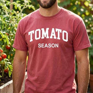 K&ouml;nnte beinhalten: Ein ziegelrotes T-Shirt mit dem wei&szlig;en Schriftzug "TOMATO SEASON". Das kurz&auml;rmelige Shirt wird von einer Person getragen. Im Hintergrund ist ein Garten mit Tomatenpflanzen zu sehen.