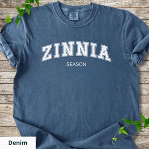 May include: Denim blue t-shirt featuring the word "ZINNIA" in large white block letters arched across the chest, with "SEASON" printed below. The shirt has a crew neck and short sleeves. The background is a wooden surface with green vines.