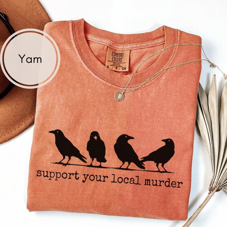 May include: A yam-colored short-sleeve t-shirt featuring a black graphic of four crows standing in a row. Below the birds, the text reads, "support your local murder." The shirt has a crew neckline and a Comfort Colors brand label inside the collar.