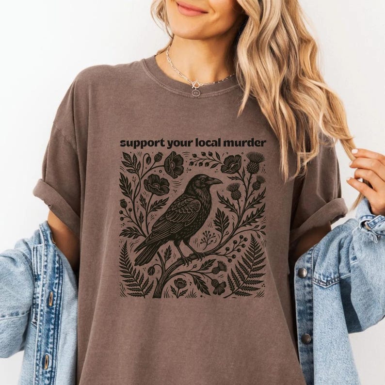 May include: A brown short-sleeved t-shirt featuring a black graphic print of a crow perched on a branch surrounded by botanical elements like flowers and ferns. The text "support your local murder" is printed in black above the illustration.
